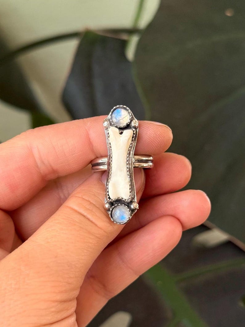 May include: A silver ring featuring a bone-shaped centerpiece with a white, textured surface. Two small, round, blue-toned gemstones are set at the top and bottom of the bone. The ring has a double band design.