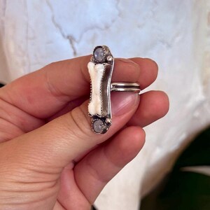 May include: A silver ring featuring a bone-shaped design with a white center and two small, round, gray stones. The ring is held in a hand, showcasing the intricate details of the jewelry. The ring's design is unique and eye-catching.
