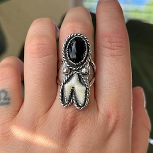 Op de afbeelding: Een zilveren ring met een zwarte ovale steen en een tandvormige witte steen. De ring heeft een gedraaid touwontwerp rond de zwarte steen en de tandvormige steen. De ring zit aan een hand.