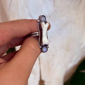 May include: A silver ring featuring a bone-shaped centerpiece with a white finish, flanked by two small, round, light blue gemstones. The ring has a detailed, textured design and is set against a blurred background.