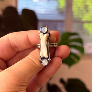 May include: A silver ring featuring a bone-shaped centerpiece with a white enamel finish. The ring is accented with two round, iridescent moonstone cabochons, one at each end of the bone design. The ring has a double band.