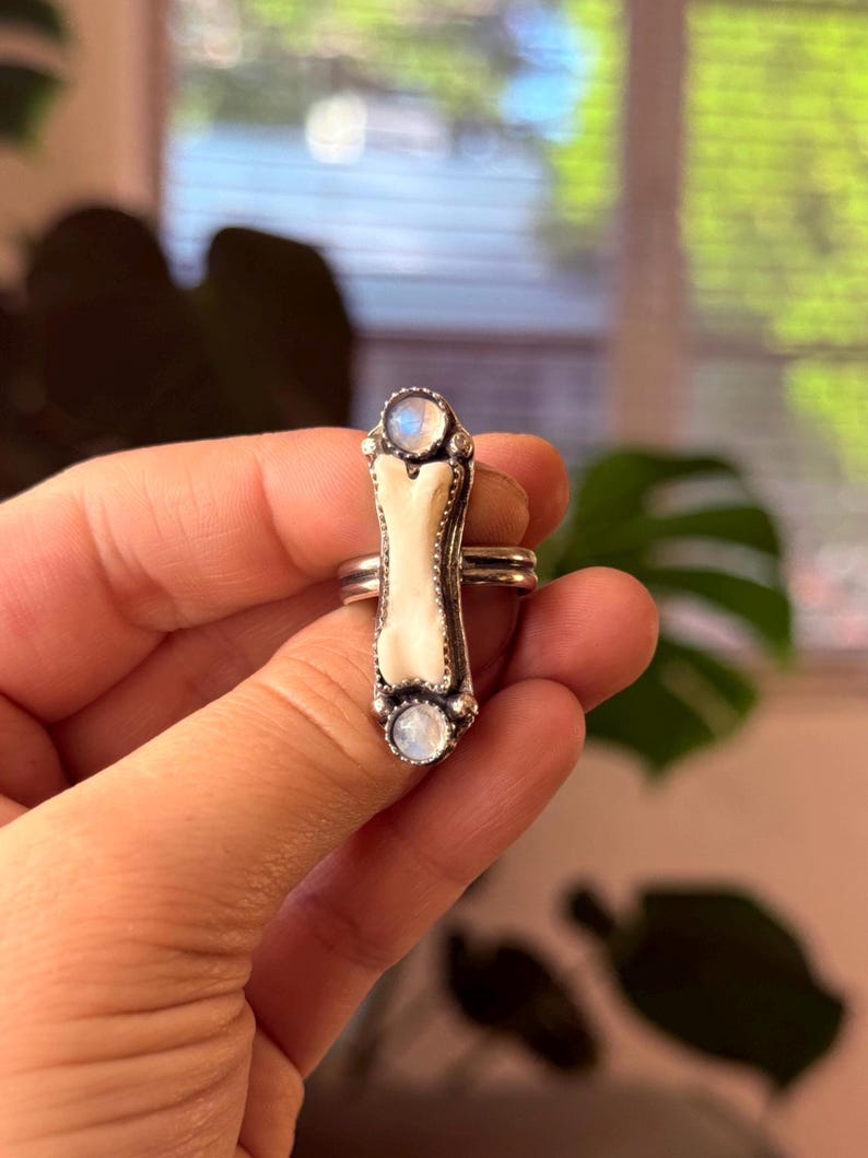 May include: A silver ring featuring a bone-shaped centerpiece with a white enamel finish. The ring is accented with two round, iridescent moonstone cabochons, one at each end of the bone design. The ring has a double band.