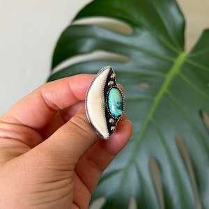 May include: A silver ring featuring a crescent-shaped white inlay alongside an oval turquoise stone with dark matrix veining. The setting includes small silver bead accents along the side of the turquoise stone and a textured silver border.