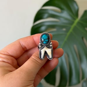 May include: A silver ring featuring an oval turquoise stone with dark matrix veining at the top, positioned above a V-shaped white inlay. The setting is framed by a twisted rope border and small silver beads, creating a detailed metal design.