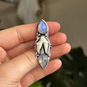 May include: A silver ring with a central tooth design, and two teardrop-shaped moonstone gems. The top gem is light blue, and the bottom gem has an iridescent quality. The ring has a unique, artistic style, suitable for alternative fashion.