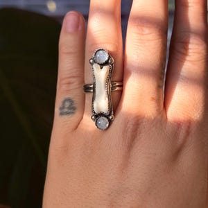 May include: A silver ring with a long, rectangular design. The ring features a central, off-white, bone-shaped element, flanked by two round, light-colored stones. The ring is worn on a finger, showcasing its intricate details and unique style.