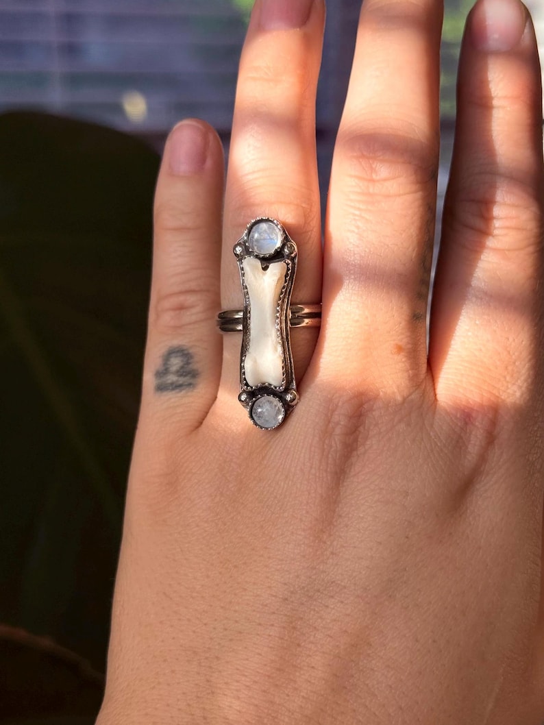 May include: A silver ring with a long, rectangular design. The ring features a central, off-white, bone-shaped element, flanked by two round, light-colored stones. The ring is worn on a finger, showcasing its intricate details and unique style.