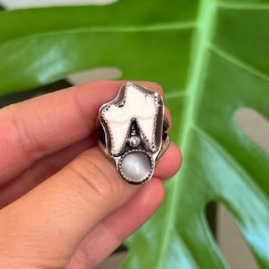 May include: A silver ring with a tooth-shaped design, featuring a white enamel finish and a small, round, opalescent stone. The ring has a detailed, textured appearance, highlighting the tooth's form and the stone's gentle sheen. A unique piece of jewellery.