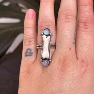 May include: A silver ring featuring a bone-shaped centerpiece with two round, light blue gemstones. The ring has a unique, handcrafted design, showcasing a blend of natural and metallic elements. The ring is worn on a finger.