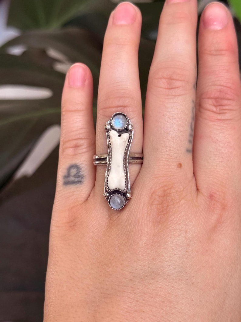 May include: A silver ring featuring a bone-shaped centerpiece with two round, light blue gemstones. The ring has a unique, handcrafted design, showcasing a blend of natural and metallic elements. The ring is worn on a finger.