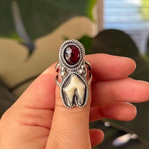 May include: A silver ring featuring a large, oval, deep red gemstone at the top and a white tooth-shaped design below. The ring has a twisted rope-like border and small gold-colored accents. The ring is held up by a hand.