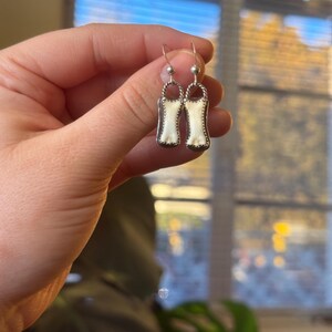 K&ouml;nnte beinhalten: Ein Paar silberne Ohrh&auml;nger. Jeder Ohrring hat einen rechteckigen, cremefarbenen Einsatz mit einer dekorativen silbernen Umrandung. Die Ohrringe sind an silbernen Haken aufgeh&auml;ngt. Das Design ist schlicht und elegant.