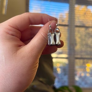K&ouml;nnte beinhalten: Silberfarbene H&auml;ngeohrringe mit rechteckigem Design und wei&szlig;-cremefarbenem, knochen&auml;hnlichem Inlay. Die Ohrringe haben einen einfachen Hakenverschluss. Sie werden in einer Hand vor unscharfem Hintergrund gehalten.