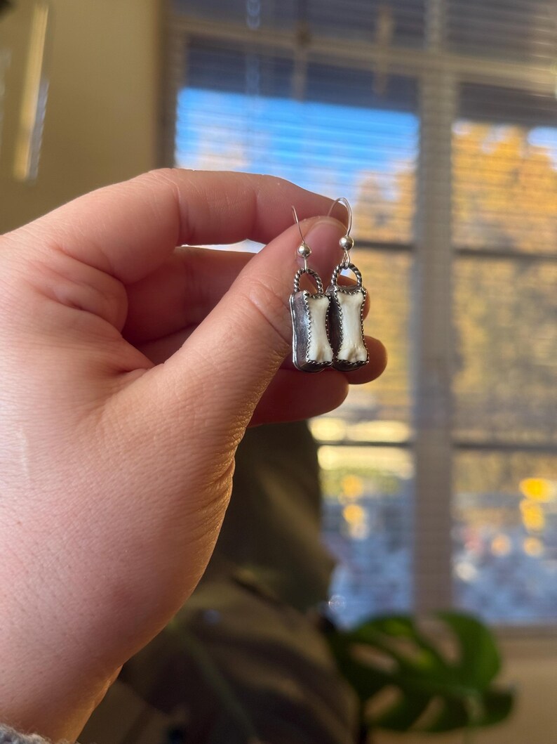 K&ouml;nnte beinhalten: Silberfarbene H&auml;ngeohrringe mit rechteckigem Design und wei&szlig;-cremefarbenem, knochen&auml;hnlichem Inlay. Die Ohrringe haben einen einfachen Hakenverschluss. Sie werden in einer Hand vor unscharfem Hintergrund gehalten.