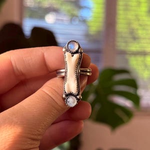 May include: A silver ring featuring a bone-shaped centerpiece with two round, iridescent moonstone accents. The ring has a detailed, ornate design and is held in a hand.