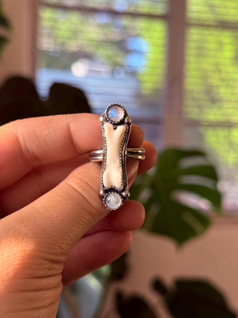 May include: A silver ring featuring a bone-shaped centerpiece with two round, iridescent moonstone accents. The ring has a detailed, ornate design and is held in a hand.