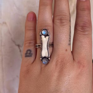 May include: A silver ring featuring a bone-shaped centerpiece with two round, light blue gemstones. The ring has a detailed, ornate design and is worn on a finger.