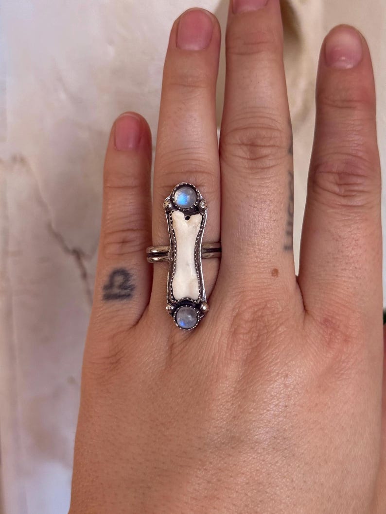 May include: A silver ring featuring a bone-shaped centerpiece with two round, light blue gemstones. The ring has a detailed, ornate design and is worn on a finger.