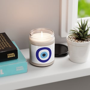 May include: A white candle with a blue and white evil eye design on the label. The candle is lit and sitting on a white shelf.