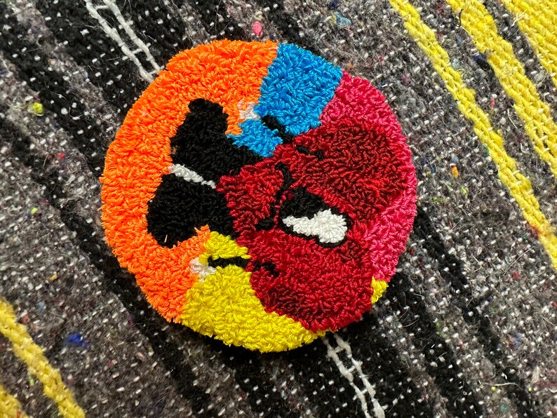 May include: A colorful, round rug with a black, white, and red design. The rug is made of yarn and has a fuzzy texture.