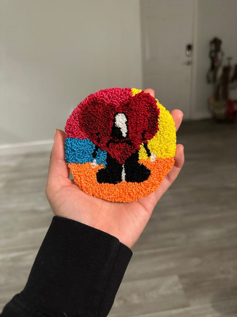 May include: A colorful round rug with a red heart in the center. The heart has a black outline and white eyes. The rug is made of yarn and has a fluffy texture.