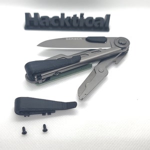 Puede incluir: Una multiherramienta gris desmontada con un mango negro y un logotipo de Gerber en la hoja. La herramienta est&aacute; sobre una superficie blanca con dos peque&ntilde;os tornillos.