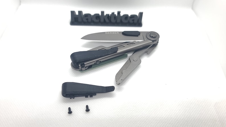 Puede incluir: Una multiherramienta gris desmontada con un mango negro y un logotipo de Gerber en la hoja. La herramienta est&aacute; sobre una superficie blanca con dos peque&ntilde;os tornillos.