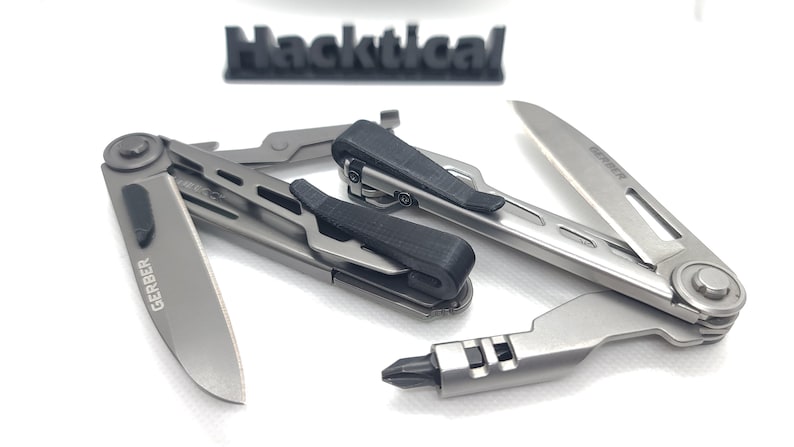 Puede incluir: Una herramienta multiusos plateada con un mango negro y una hoja que dice "GERBER". La herramienta est&aacute; abierta y la hoja est&aacute; extendida. La herramienta est&aacute; sobre una superficie blanca.