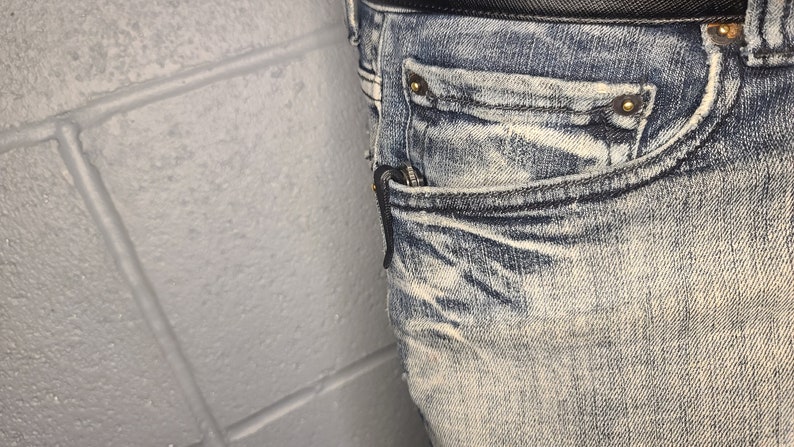 Puede incluir: Primer plano de un bolsillo de mezclilla azul claro con una cremallera negra y remaches dorados. La mezclilla tiene un aspecto descolorido y desgastado.