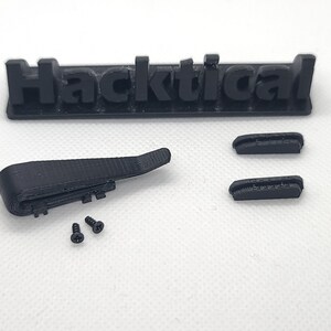 Puede incluir: Una pieza impresa en 3D de color negro con dos tornillos negros. La pieza tiene forma de pieza plana curva con una peque&ntilde;a pieza rectangular unida a ella. La pieza est&aacute; etiquetada como "Hacktical".