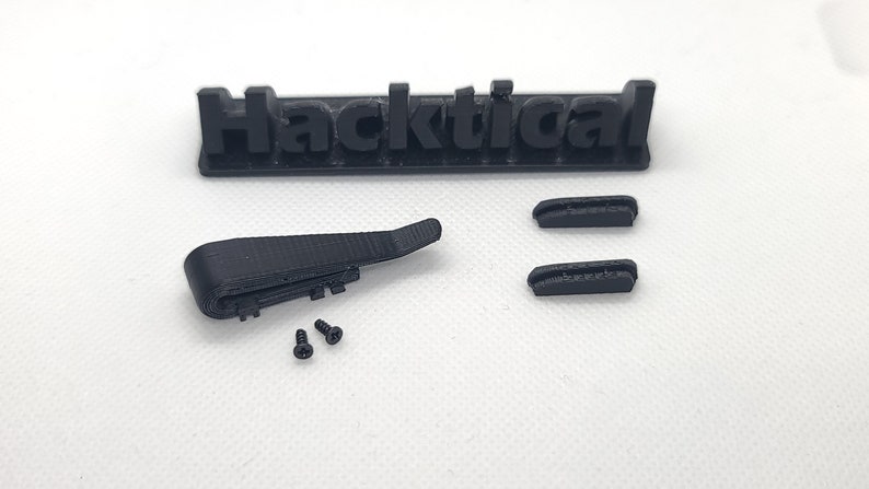 Puede incluir: Una pieza impresa en 3D de color negro con dos tornillos negros. La pieza tiene forma de pieza plana curva con una peque&ntilde;a pieza rectangular unida a ella. La pieza est&aacute; etiquetada como "Hacktical".