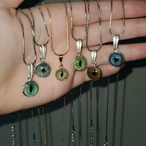 May include: A selection of eye pendant necklaces in shades of blue, green, and brown. The round pendants are set in silver or gold-coloured metal. The necklaces are shown against a plain backdrop.