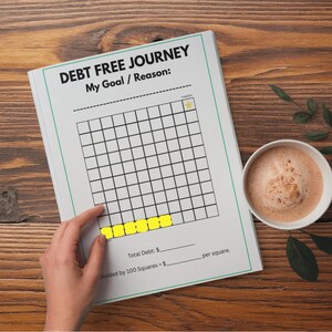 Puede incluir: Un cuaderno blanco con el texto "DEBT FREE JOURNEY" y una cuadrícula para el seguimiento de objetivos financieros. Una mano señala los cuadrados amarillos resaltados. Una taza de café está cerca sobre una superficie de madera.