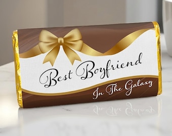 May include: A rectangular chocolate bar wrapper featuring a brown and white color scheme with a gold ribbon bow graphic. The text "Best Boyfriend" is printed in a black script font, followed by "In The Galaxy" in a smaller white font.