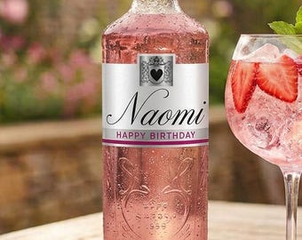 May include: A clear glass bottle filled with a pink liquid, featuring a white label with the text Naomi and HAPPY BIRTHDAY, alongside a stemmed glass containing a pink cocktail garnished with fresh strawberry slices and a green mint sprig.