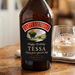 May include: A dark brown glass bottle with a custom black label featuring the text Happy Birthday, a landscape illustration, the name TESSA, and the message Enjoy your special day FROM LUCY XX, accompanied by a clear glass tumbler containing a beige drink.