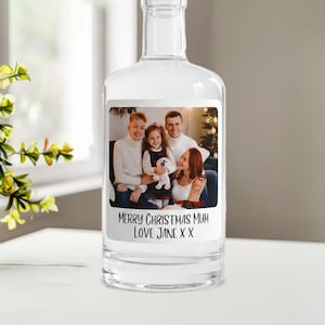 May include: A clear glass bottle featuring a rectangular label with a photograph of a family of four. The label includes the printed text: MERRY CHRISTMAS MUM LOVE JANE X X. The bottle has a narrow neck and a smooth, transparent glass body.