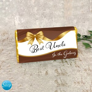 May include: A rectangular chocolate bar wrapper featuring a dark brown background with a white center panel, a gold ribbon bow graphic, and the black text "Best Uncle In the Galaxy." The ends of the wrapper are finished in shiny gold foil.