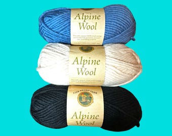 May include: Three skeins of Alpine Wool yarn in blue, white, and black. Each skein has a paper label with the text "Alpine Wool" and product information. The yarn is soft and natural, suitable for various crafting projects.