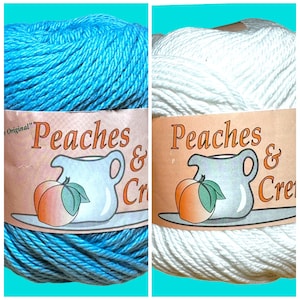 May include: Two skeins of Peaches & Creme cotton yarn, one in turquoise and the other in white. Each skein has a label featuring a peach, a pitcher, and the words "Peaches & Creme". The yarn is tightly wound.