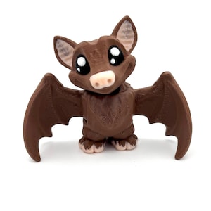 May include: A brown bat figurine with large, black eyes and a pink nose. The bat has brown wings and is standing on a white surface.