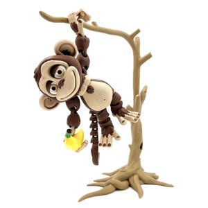 May include: A brown monkey figurine with a yellow banana hanging from its hand, hanging from a brown tree branch. The monkey has a smiling face and is posed as if it is swinging from the branch.