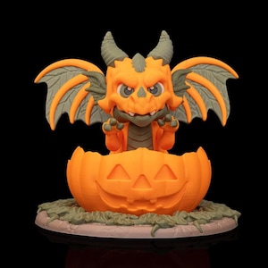 May include: A small orange and olive green dragon figurine emerging from a carved orange jack-o'-lantern pumpkin. The dragon has wings, horns, and claws, and is positioned on a circular base with textured green details resembling foliage.