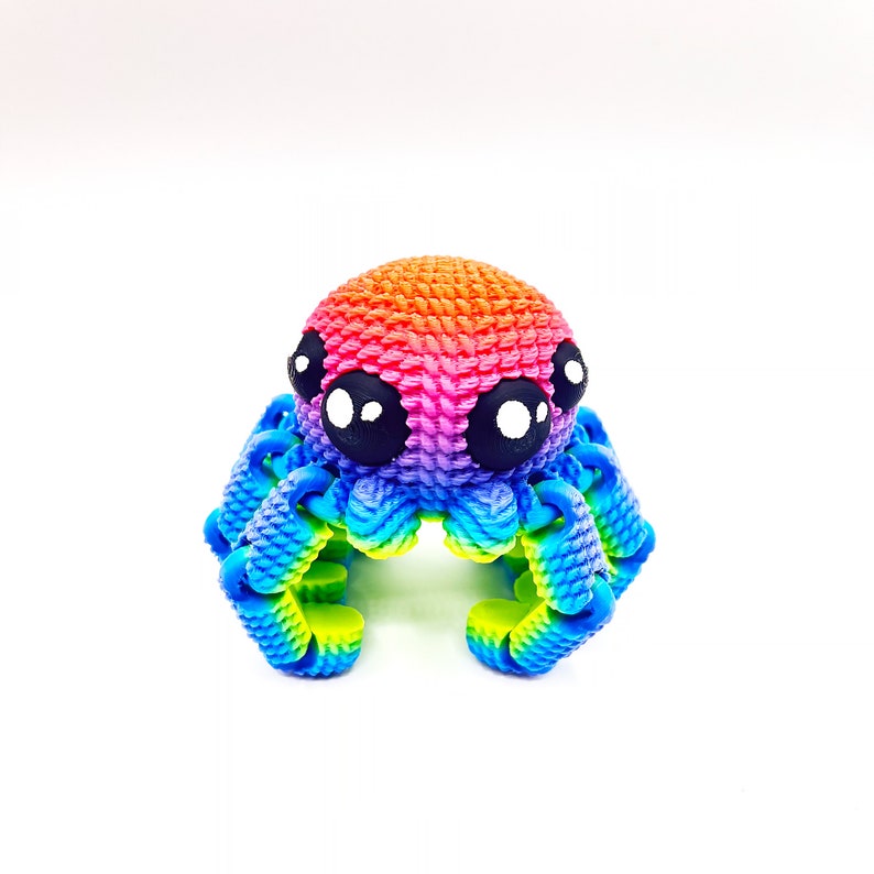 Op de afbeelding: Een kleine, regenboogkleurige gehaakte octopus met grote zwarte ogen en witte pupillen. De octopus heeft acht tentakels en is gemaakt van een zacht, getextureerd garen.