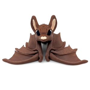 May include: A brown bat figurine with large black eyes and a pink nose. The bat has its wings spread out, and it is made of a textured material.