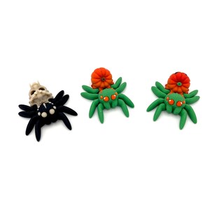 May include: Three Halloween-themed spider figurines. One black spider with a skull on its back, and two green spiders with orange pumpkin accents. The spiders have orange eyes and are set against a white background.