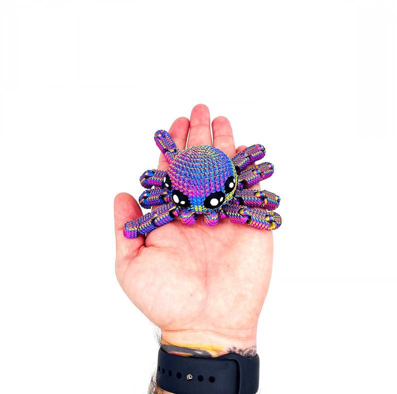 Op de afbeelding: Een kleine, regenboogkleurige, gehaakte spin met zwarte ogen. De spin is roze, blauw en groen.