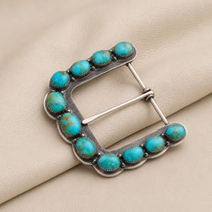 May include: A rectangular belt buckle crafted from dark oxidised metal, featuring a scalloped edge border. The frame is inlaid with ten oval-shaped turquoise stones, each displaying unique brown matrix patterns and held by a serrated bezel setting.