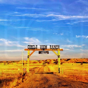 May include: A wooden sign with the text "Circle View Ranch" above a dirt road leading into a field of dry grass. The sky is blue with white clouds.