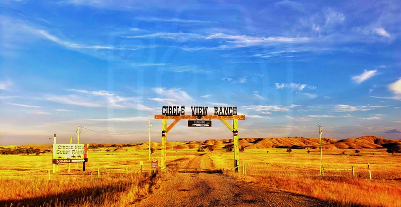 May include: A wooden sign with the text "Circle View Ranch" above a dirt road leading into a field of dry grass. The sky is blue with white clouds.